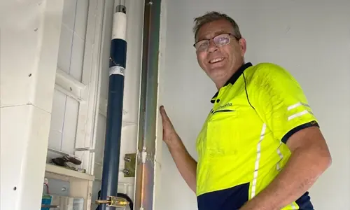 Smiling elevator technician wearing a high‑visibility safety shirt, standing beside residential elevator piping and mechanical components during lift installation or service.