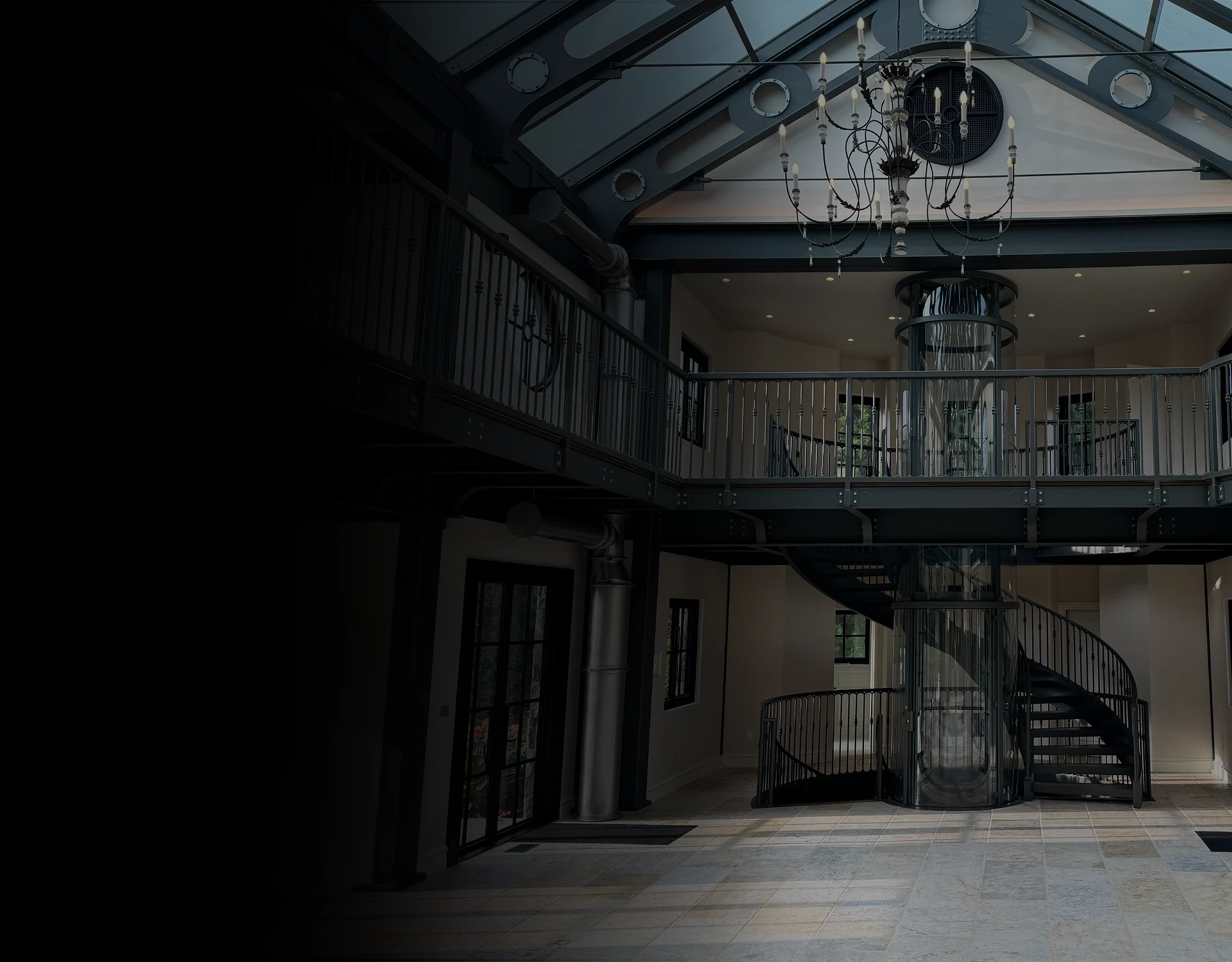 Glass cylindrical elevator in a grand atrium with chandelier lighting, mezzanine railings, and a spiral staircase