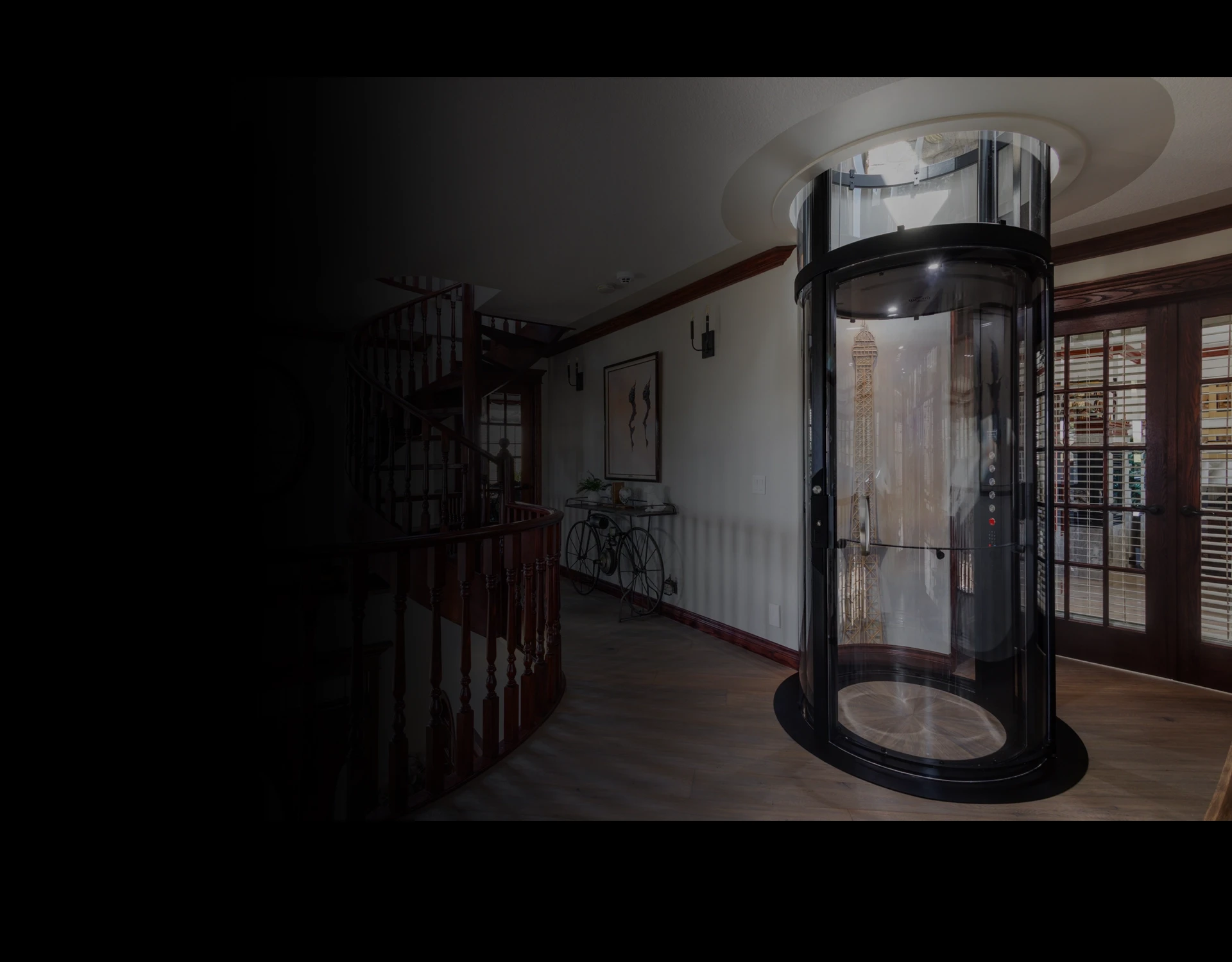 Glass cylindrical residential elevator in a grand foyer beside a curved staircase and wood flooring